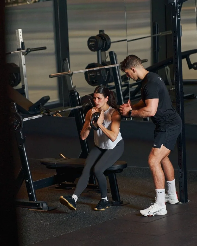 Personal trainer che supervisiona un’atleta nello squat con kettlebell in una palestra luminosa.